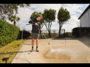 Person using a pressure washer to clean a concrete surface in a residential outdoor setting, surrounded by greenery and a fence, illustrating pressure washing services offered by Spray Down Exterior Cleaning in Sawgrass, FL.