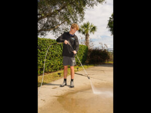Young man using a pressure washer on a concrete surface, demonstrating exterior cleaning services in a residential setting with greenery in the background, relevant to Spray Down Exterior Cleaning's offerings in Mandarin, FL.