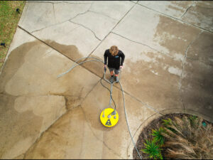 Person using a power washer on a concrete sidewalk, showcasing effective commercial cleaning services for storefronts, with visible dirt removal and a clean, manicured appearance.